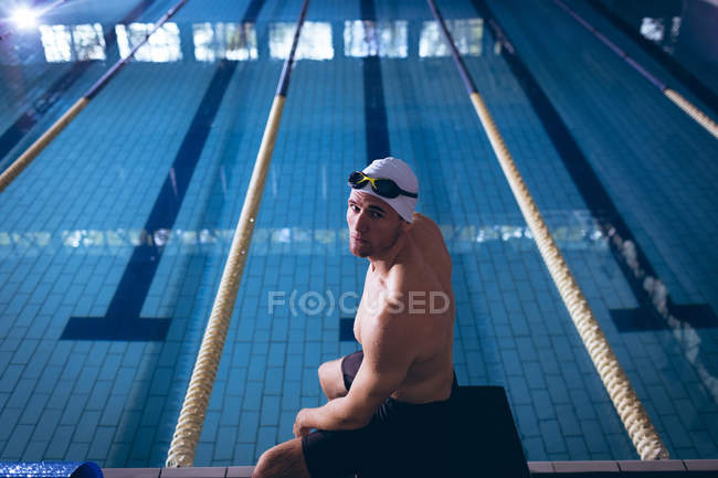 Vista lateral de un nadador caucásico macho sentado en un trampolín junto a la piscina - foto de stock
