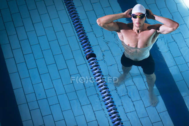 Vue en angle élevé d'un homme nageur caucasien portant un bonnet de bain blanc avec des lunettes tout en se tenant dans la piscine — Photo de stock
