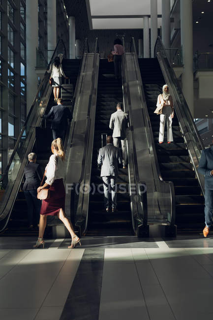 Vista frontal de diversos empresarios que utilizan escaleras mecánicas en oficinas modernas - foto de stock