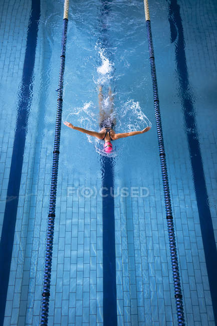 Vista de alto ângulo de uma mulher caucasiana vestindo um maiô e um boné de natação rosa fazendo um derrame de borboleta na piscina — Fotografia de Stock