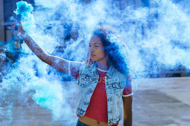 Vista frontal de uma jovem afro-americana vestindo um colete de ganga segurando um fabricante de fumaça produzindo fumaça azul — Fotografia de Stock