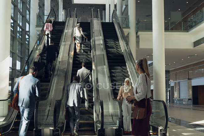Вид спереди различных бизнесменов, использующих эскалаторы в современном офисе — стоковое фото
