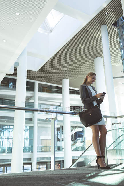 Vista de ángulo bajo de la mujer de negocios apoyada en la barandilla y el uso de teléfono móvil en el moderno edificio de oficinas - foto de stock