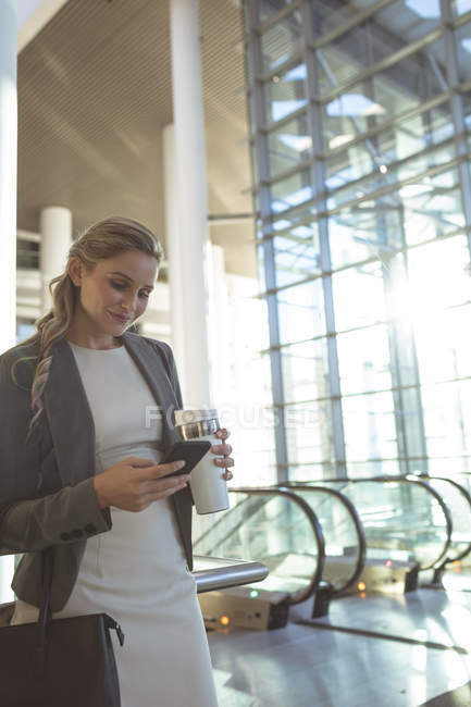 Vista lateral da bela mulher de negócios usando telefone celular e segurando xícara de café descartável em um edifício de escritório moderno — Fotografia de Stock