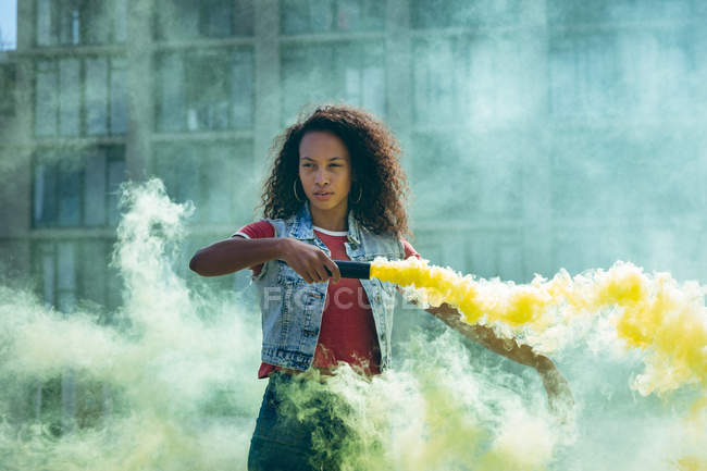 Vista frontal de una joven afroamericana vestida con un chaleco vaquero sosteniendo una máquina de humo que produce humo amarillo en una azotea con vista a un edificio y la luz del sol - foto de stock