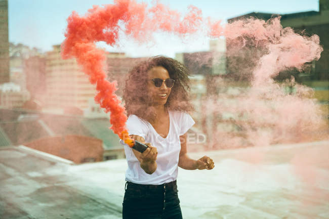 Vista frontal de una joven afroamericana con una camisa blanca y anteojos sonriendo mientras sostiene una máquina de humo que produce humo naranja en una azotea con vistas a edificios - foto de stock