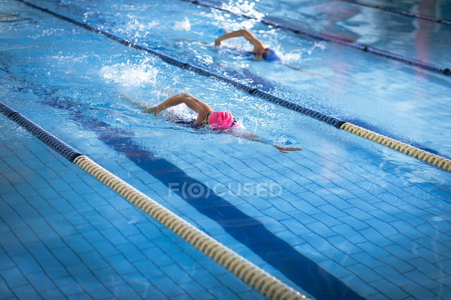 Seitenansicht einer jungen afrikanisch-amerikanischen und kaukasischen Frau beim Freestyle-Schlaganfall im Pool, während die Schwimmerin mit pinkfarbener Mütze führt — Stockfoto