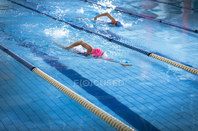 Vista lateral de uma jovem afro-americana e caucasiana fazendo freestyle acidente vascular cerebral na piscina enquanto o nadador com boné rosa leva — Fotografia de Stock