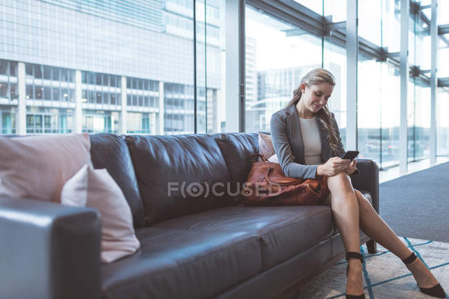 Side view of beautiful businesswoman using mobile phone on sofa in the lobby at modern office — Stock Photo
