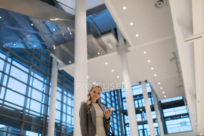 Низкий угол обзора счастливой деловой женщины, смотрящей в камеру, используя мобильный телефон в вестибюле современного офиса — стоковое фото