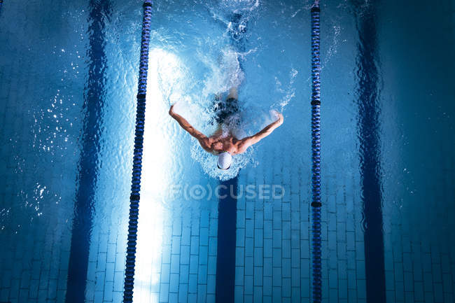 Vista de ángulo alto de un nadador caucásico masculino con una gorra de natación blanca haciendo un golpe de mariposa en la piscina - foto de stock