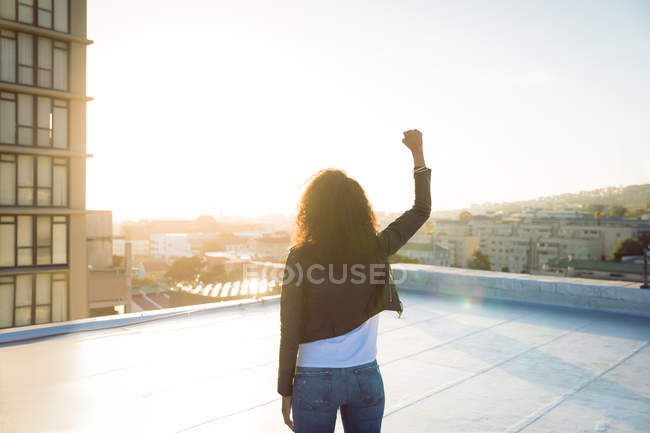 Vista posteriore di una giovane donna afro-americana che indossa una giacca di pelle con pugno alzato mentre si trova su un tetto con vista su un edificio e la luce del sole — Foto stock