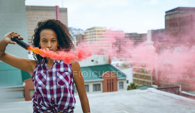 Вид спереди на молодую афроамериканку в клетчатом топе, держащую дымовую трубу, производящую красный дым на крыше с видом на здания, при этом пристально глядя в камеру — стоковое фото