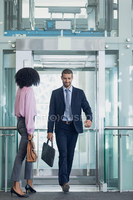 Vista frontal del ejecutivo masculino caucásico saliendo del ascensor en la oficina moderna. Mujer afroamericana esperando para entrar en el ascensor - foto de stock