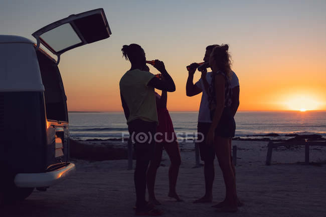 Vista lateral do grupo de diversos amigos bebendo cerveja perto de van campista durante o pôr do sol — Fotografia de Stock