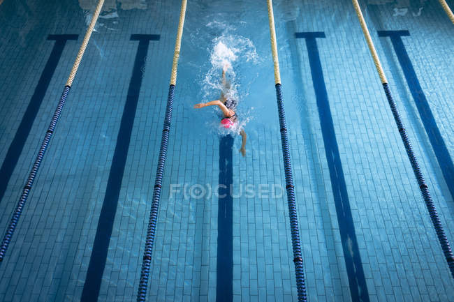 Высокий угол обзора белой женщины в купальнике и розовой плавательной шапке, делающей фристайл-инсульт в бассейне — стоковое фото