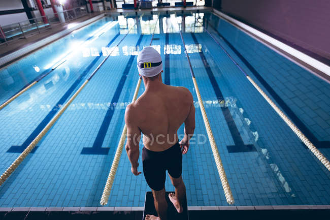 Rear view of a male Caucasian swimmer wearing a white swimming cap standing by an olympic sized pool inside a stadium — Stock Photo