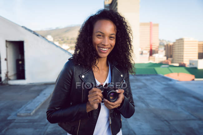 Vorderseite einer jungen afrikanisch-amerikanischen Frau, die eine Lederjacke trägt und lächelt, während sie eine Kamera auf einem Dach hält — Stockfoto