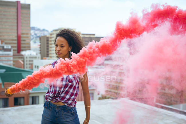 Frontansicht einer jungen afrikanisch-amerikanischen Frau, die ein kariertes Oberteil trägt, während sie eine Rauchmaschine hält, die roten Rauch auf einem Dach mit Blick auf Gebäude produziert — Stockfoto