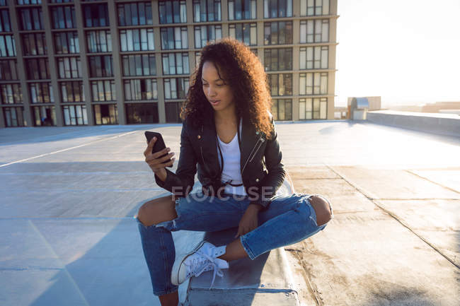Vista frontale di una giovane donna afro-americana che indossa una giacca di pelle seduta e utilizza un telefono cellulare su un tetto con vista su un edificio e sulla luce del sole — Foto stock