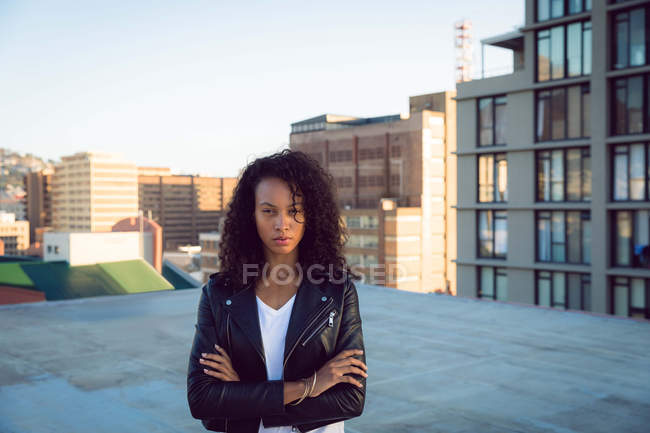 Вид спереди на молодую афроамериканку в кожаной куртке, пристально смотрящую на камеру со скрещенными руками, стоящую на крыше с видом на здания — стоковое фото