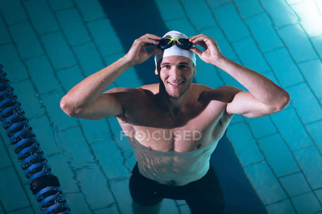Hochwinkelaufnahme eines männlichen kaukasischen Schwimmers mit weißer Badekappe, der lächelnd die Brille im Kopf hält, während er im Schwimmbad steht — Stockfoto