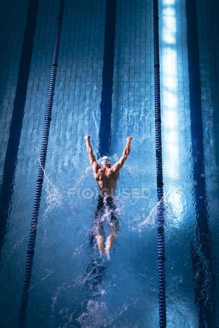 Vista de ángulo alto de un nadador caucásico masculino con una gorra de natación blanca haciendo un golpe de mariposa en la piscina - foto de stock
