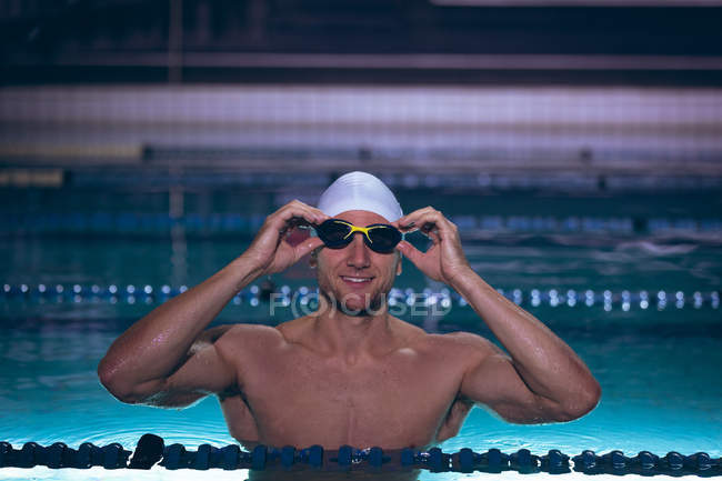 Vorderansicht eines männlichen kaukasischen Schwimmers, der die Brille auf seiner weißen Schwimmmütze hält, während er im Schwimmbad steht — Stockfoto