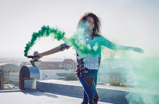 Vista frontal de uma jovem afro-americana vestindo um colete de ganga segurando um fumante produzindo fumaça verde em um telhado com luz solar — Fotografia de Stock