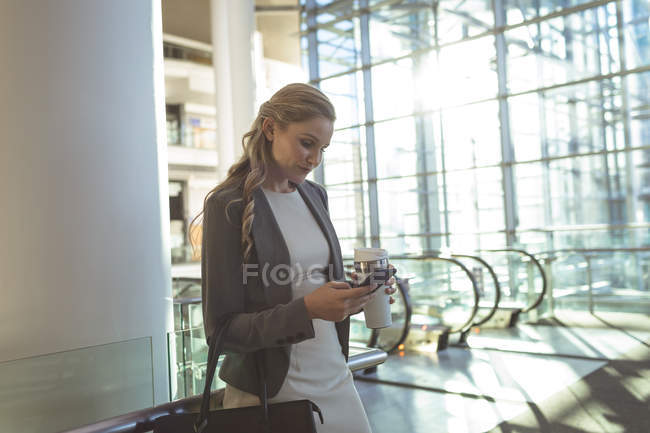Vista lateral de la hermosa mujer de negocios utilizando el teléfono móvil y la celebración de la taza de café desechable en un moderno edificio de oficinas - foto de stock