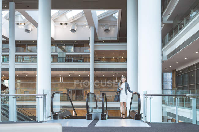 Vista frontal de empresária usando telefone celular na escada rolante em um prédio de escritórios moderno — Fotografia de Stock