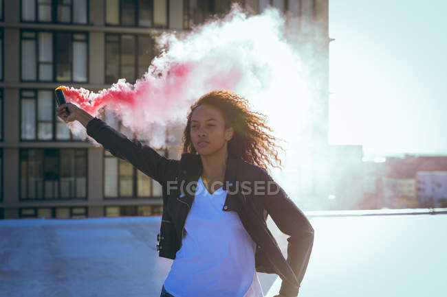 Vista frontal de uma jovem afro-americana vestindo uma jaqueta de couro segurando um fabricante de fumaça produzindo fumaça vermelha em um telhado com vista para um prédio e luz solar — Fotografia de Stock
