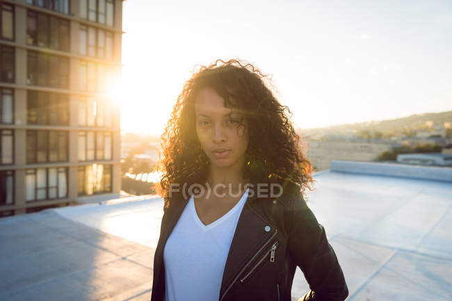 Vista frontale di una giovane donna afro-americana che indossa una giacca di pelle guardando attentamente la telecamera mentre si trova su un tetto con vista su un edificio e sul tramonto — Foto stock
