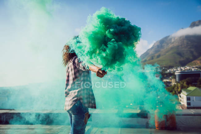Side view of a young African-American woman wearing a plaid jacket holding a smoke maker producing green smoke on a rooftop — Stock Photo