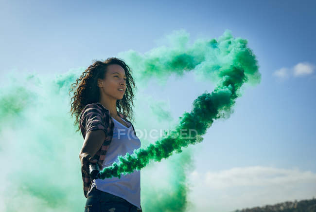 Side view of a young African-American woman wearing a plaid jacket holding a smoke maker producing green smoke on a rooftop — Stock Photo
