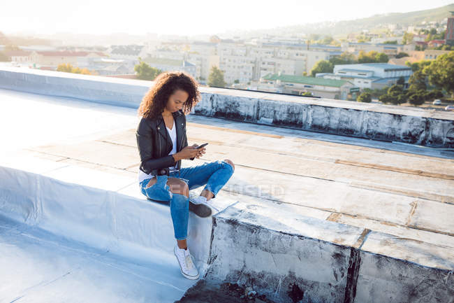 Vista laterale di una giovane donna afro-americana che indossa una giacca di pelle seduta e utilizza un telefono cellulare su un tetto — Foto stock