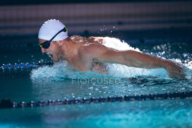 Vue latérale d'un homme nageur caucasien portant un bonnet de bain blanc et des lunettes faisant un coup de papillon dans la piscine — Photo de stock