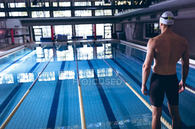 Заднього виду чоловічого Кавказького плавця в білому ковпачком стояв Олімпійський розмір басейну всередині стадіону — стокове фото