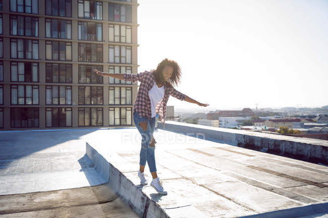 Front view of a young African-American woman wearing a plaid jacket with hands stretched while walking on a rooftop with a view of a building and sunlight — Stock Photo
