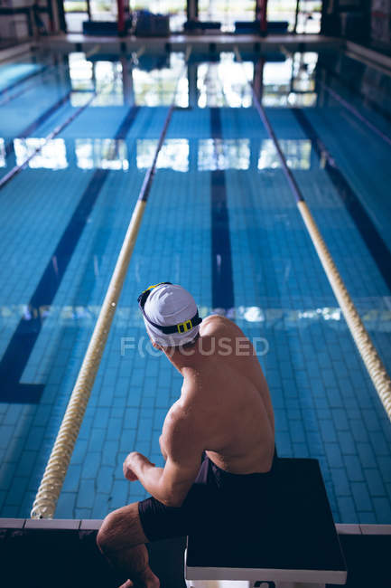 Rückansicht eines männlichen kaukasischen Schwimmers, der auf einem Sprungbrett neben dem Schwimmbad sitzt — Stockfoto