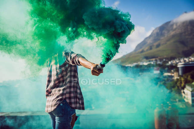 Vista lateral de una joven afroamericana con una chaqueta a cuadros sosteniendo una máquina de humo que produce humo verde en una azotea - foto de stock