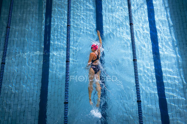 Vista de ángulo alto de una mujer caucásica con una gorra de natación rosa y gafas haciendo un golpe de estilo libre en una piscina - foto de stock