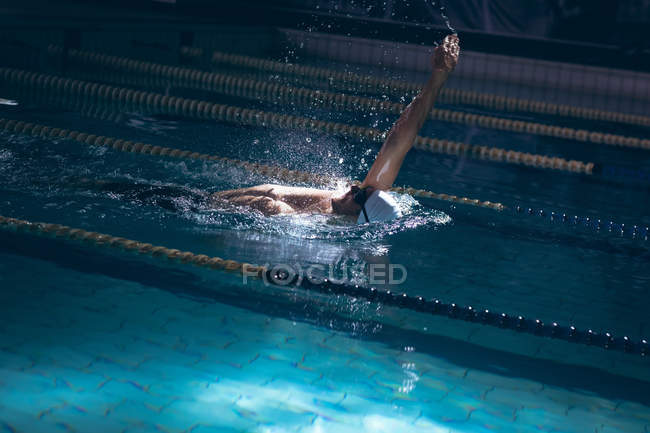 Side view of a male Caucasian swimmer wearing a white swimming cap and goggles doing a back stroke in the swimming pool — Stock Photo