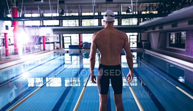 Vista posteriore di un nuotatore caucasico di sesso maschile che indossa un cappello da bagno bianco in piedi vicino a una piscina olimpionica all'interno di uno stadio — Foto stock