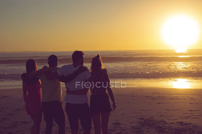 Вид ззаду різних друзів, що стоять з обіймами на пляжі під час заходу сонця — стокове фото