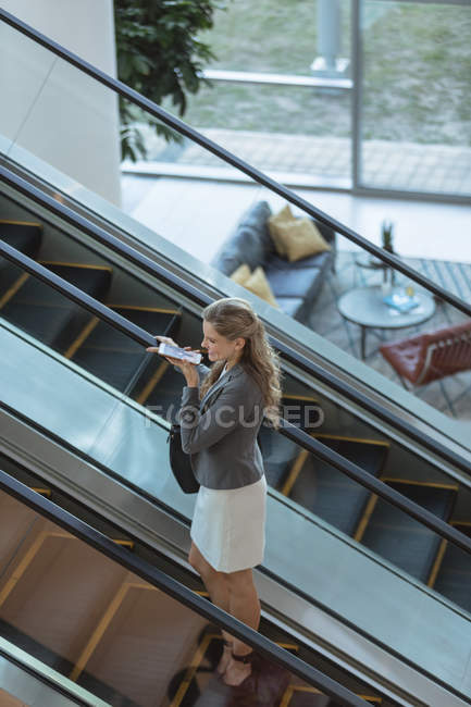 Hochwinkelaufnahme einer Geschäftsfrau, die auf einer Rolltreppe in einem modernen Bürogebäude mit dem Handy spricht — Stockfoto