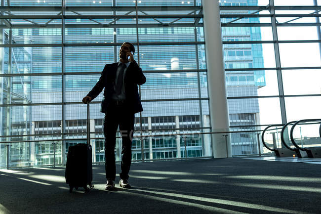 Frontansicht eines Geschäftsmannes mit Rollkoffer im Gespräch mit dem Handy im modernen Bürogebäude — Stockfoto