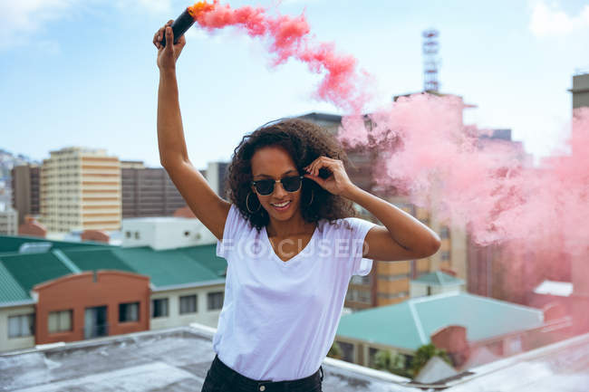 Vista frontale di una giovane donna afro-americana che indossa una camicia bianca e un occhiale sorridente mentre tiene in mano un fumatore che produce fumo rosso su un tetto con vista sugli edifici — Foto stock