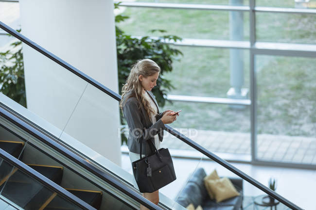 Vista lateral de empresária usando telefone celular na escada rolante em um prédio de escritórios moderno — Fotografia de Stock