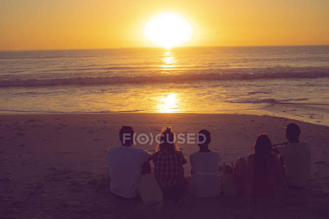 Rear view of diverse friends sitting together on the beach during sunset — Stock Photo